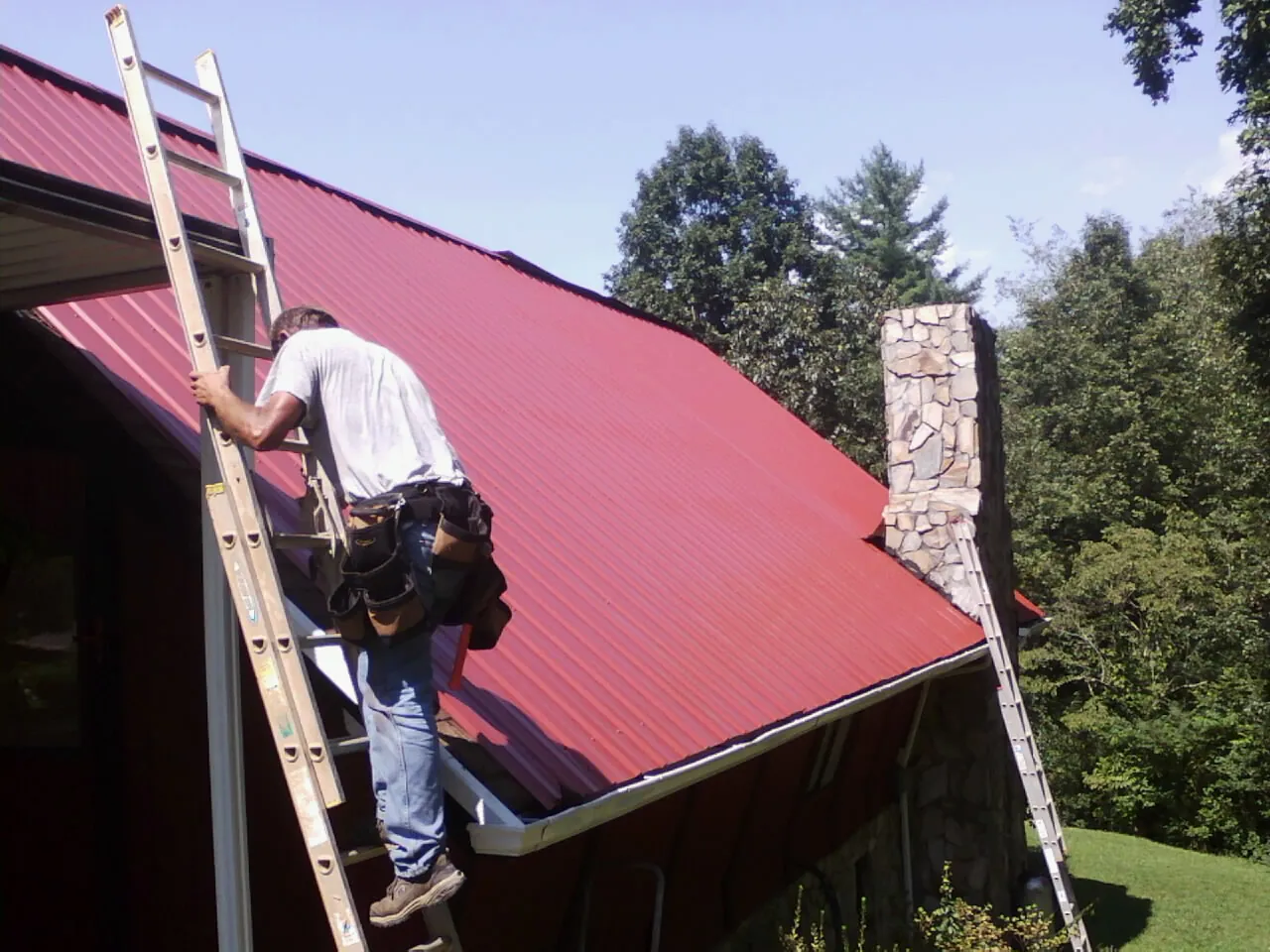 Roofer on a ladder beside a red metal roof and stone chimney — Soffit & Fascia Repair by Roofing Brothers in Adkins